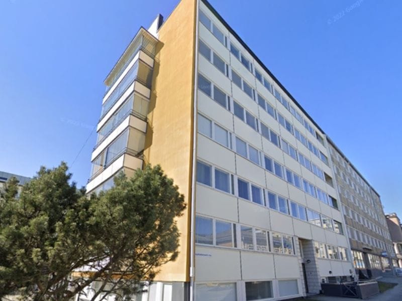 Vaalea kerrostalo, korkea, vaalea rakennus, jossa parvekkeita ja ikkunoita, taustalla sininen taivas ja etualalla vihreä nurmikko puineen