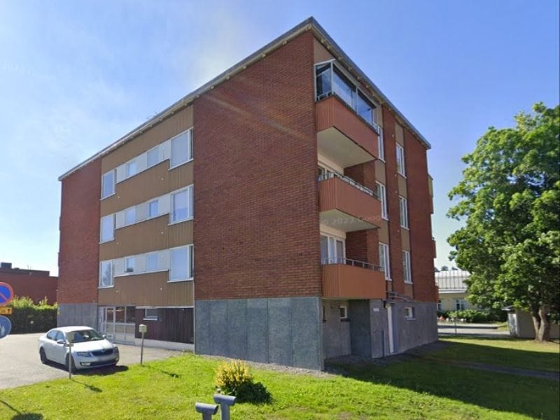  As Oy Jormankulma Nilsiässä (Kuopio), nelikerroksinen punatiilinen rakennus, jossa on parvekkeita ja harmaa kivijalka. Edessä näkyy parkkipaikalla pysäköity auto ja taustalla puu vehreällä nurmikolla