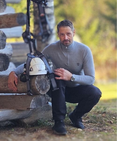 Yrityksen omistaja Juhani kyykyssä hirsirakennuksen kulmassa, metsä taustalla, kädessään kypärä ja turvavaljaat, jotka roikkuvat puuhun kiinnitetyssä koukussa.