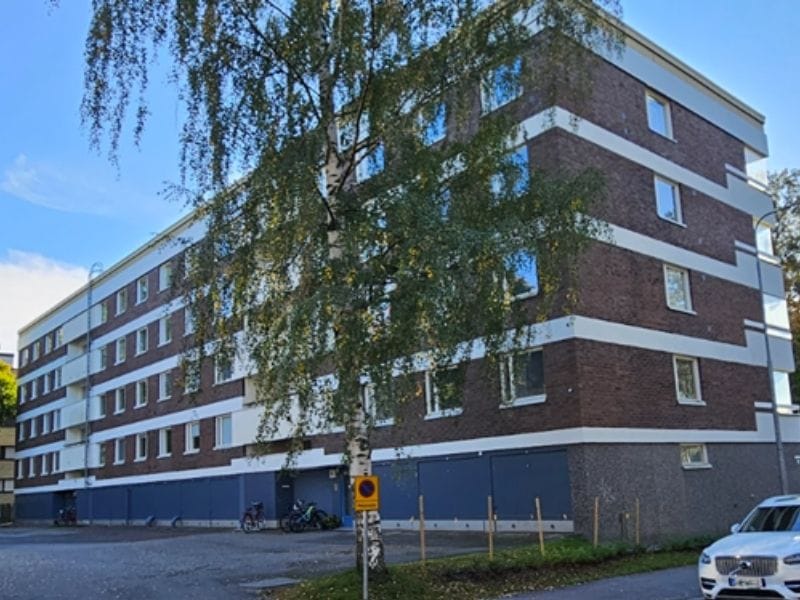 Kallanranta (800 x 600 px) Nelikerroksinen kerrostalo, jossa on tummanruskea tiilijulkisivu ja valkoisia vaakaraitoja, etualalla lehtipuu ja pysäköity auto kirkkaassa päivänvalossa