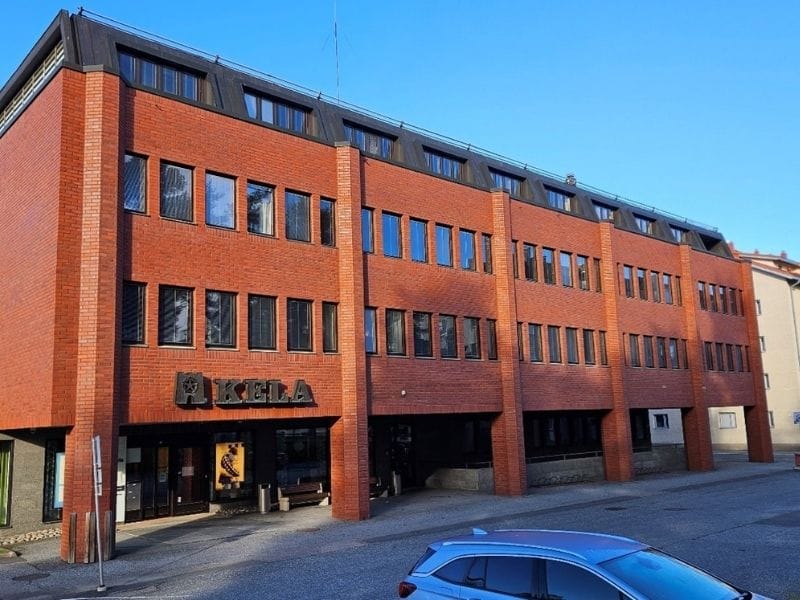 Tiilinen kolmikerroksinen rakennus Kelan toimisto, moderni punatiilinen julkisivu ja iso Kela-logo, etualalla pysäköity auto kirkkaassa päivänvalossa