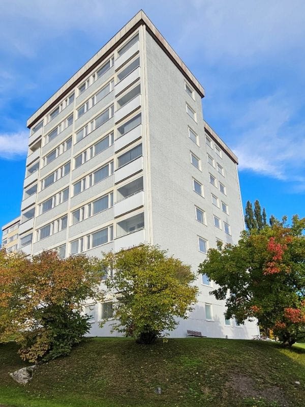 Vaalea kerrostalo, jossa on kahdeksan kerrosta ja lasitetut parvekkeet, ympärillä värikkäitä puita ja vihreä nurmikko, taustalla kirkas sininen taivas.