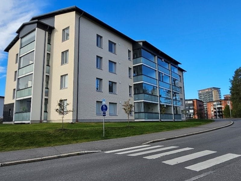 Nelikerroksinen vaalea kerrostalo, suuria parvekeikkunoita ja vihreä nurmikko talon edustalla, kirkas sininen taivas taustalla.