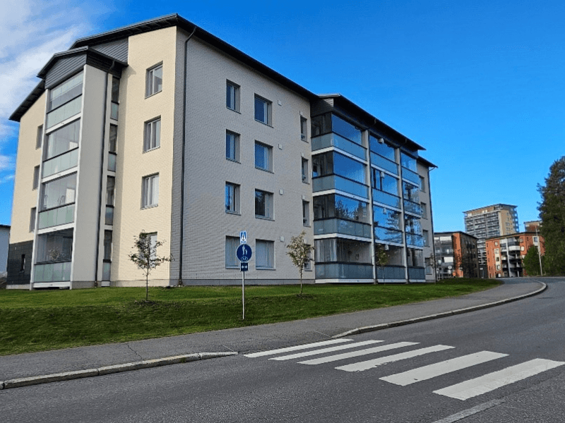 Nelikerroksinen vaalea kerrostalo, suuria parvekeikkunoita ja vihreä nurmikko talon edustalla, kirkas sininen taivas taustalla.