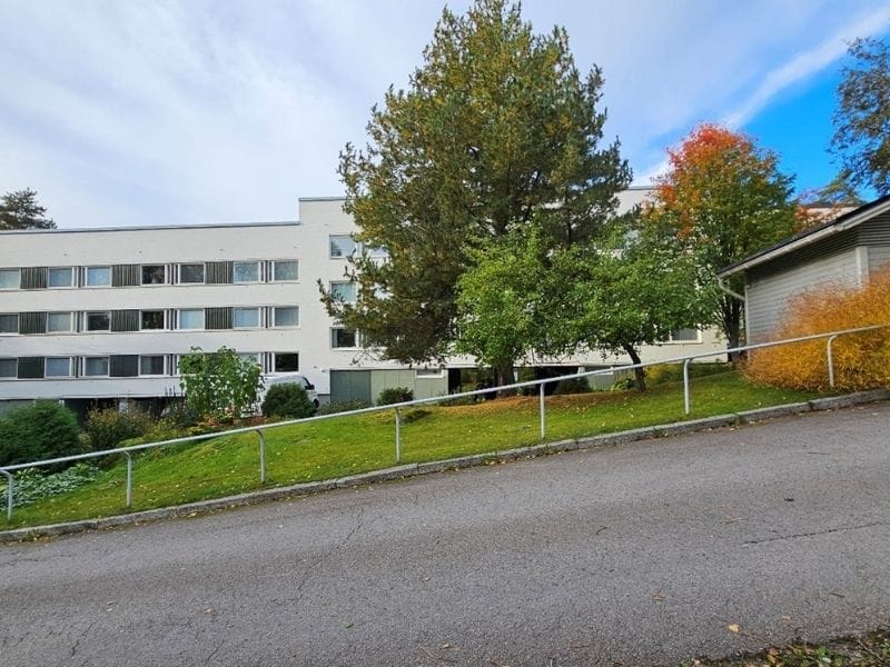 Vaalea kolmikerroksinen kerrostalo Luistelijantiellä, edessä pieni mäkinen piha-alue ja vaalea aita, ympärillä puita ja vihreää nurmikkoa.