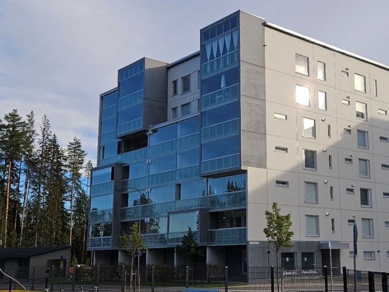 Moderni kerrostalo, jossa suuria lasiparvekkeita ja harmaata julkisivua, rakennus seisoo metsän reunalla sinistä taivasta vasten