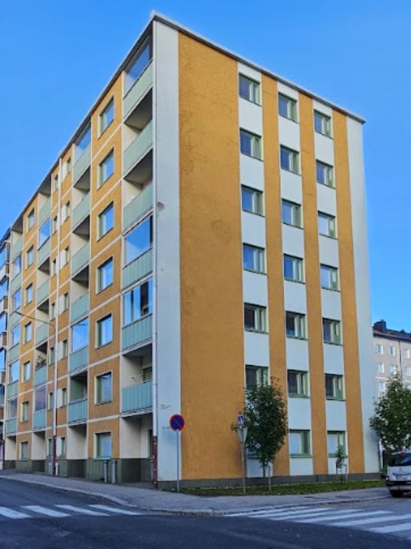 Oranssi ja vaalea kerrostalo Mäkikadulla, kuusi kerrosta, parvekkeita ja ikkunarivejä, edessä suojatie ja pienet puut kadun varrella