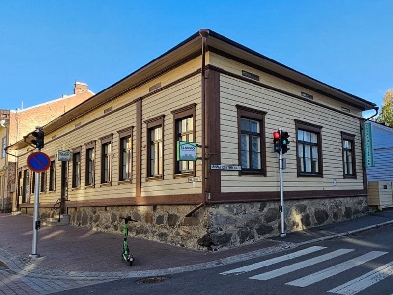 Vaalea puinen matala rakennus, jossa on ruskeat kulmalistat ja kivijalka, nurkalla liikennevalot ja sähköpotkulauta kadun laidalla.