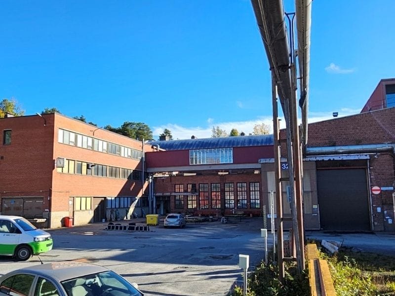 VR-Konepaja (800 x 600 px) Punatiilinen teollisuusrakennus, suuri piha-alue, putkia ja pysäköityjä autoja, taustalla sininen taivas ja varjoisa alue