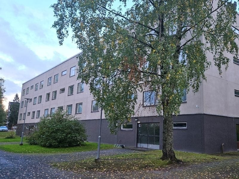 Nelikerroksinen vaalea kerrostalo, edessä lehtipuu ja sorapäällysteinen piha, puut osittain varjostavat rakennuksen julkisivua