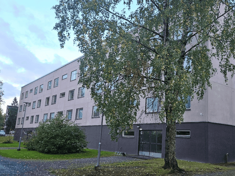 Vuorelan_Possu Nelikerroksinen vaalea kerrostalo, edessä lehtipuu ja sorapäällysteinen piha, puut osittain varjostavat rakennuksen julkisivua